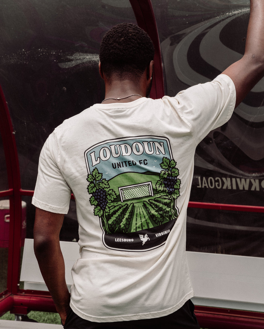 Person wearing a white t-shirt with a graphic design on the back, standing next to a vehicle.