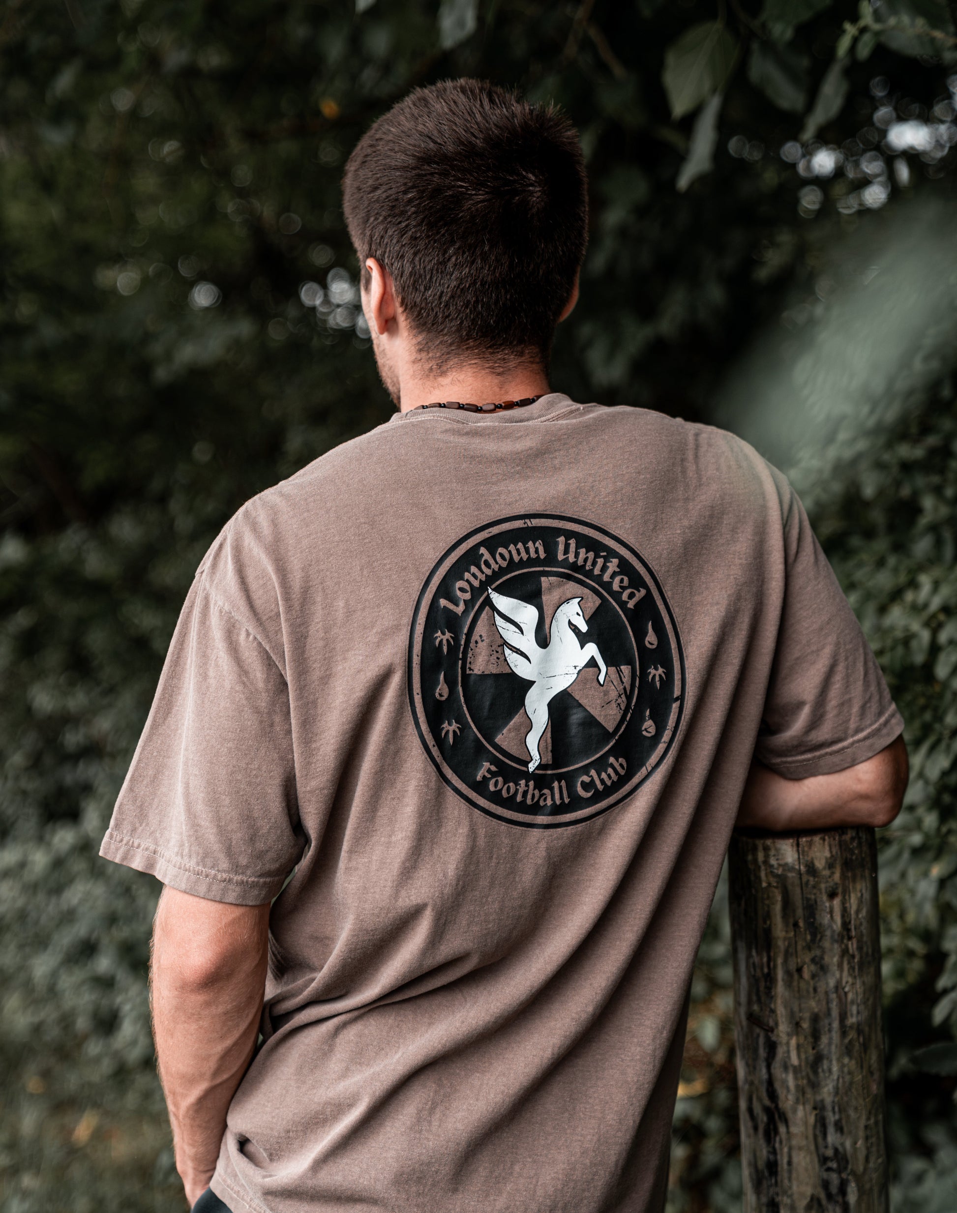 A man viewed from behind wearing a brownish-gray t-shirt with a large circular London United Football Club graphic on the back, standing outdoors next to a wooden post with green foliage in the background.