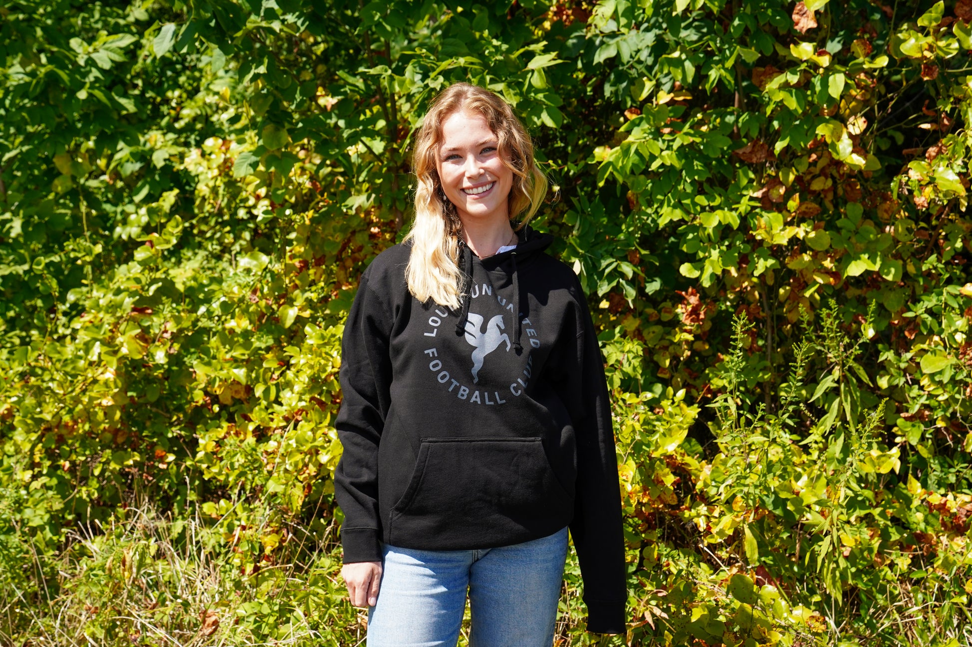 Woman wearing a black hoodie with a graphic design, standing in front of green foliage.