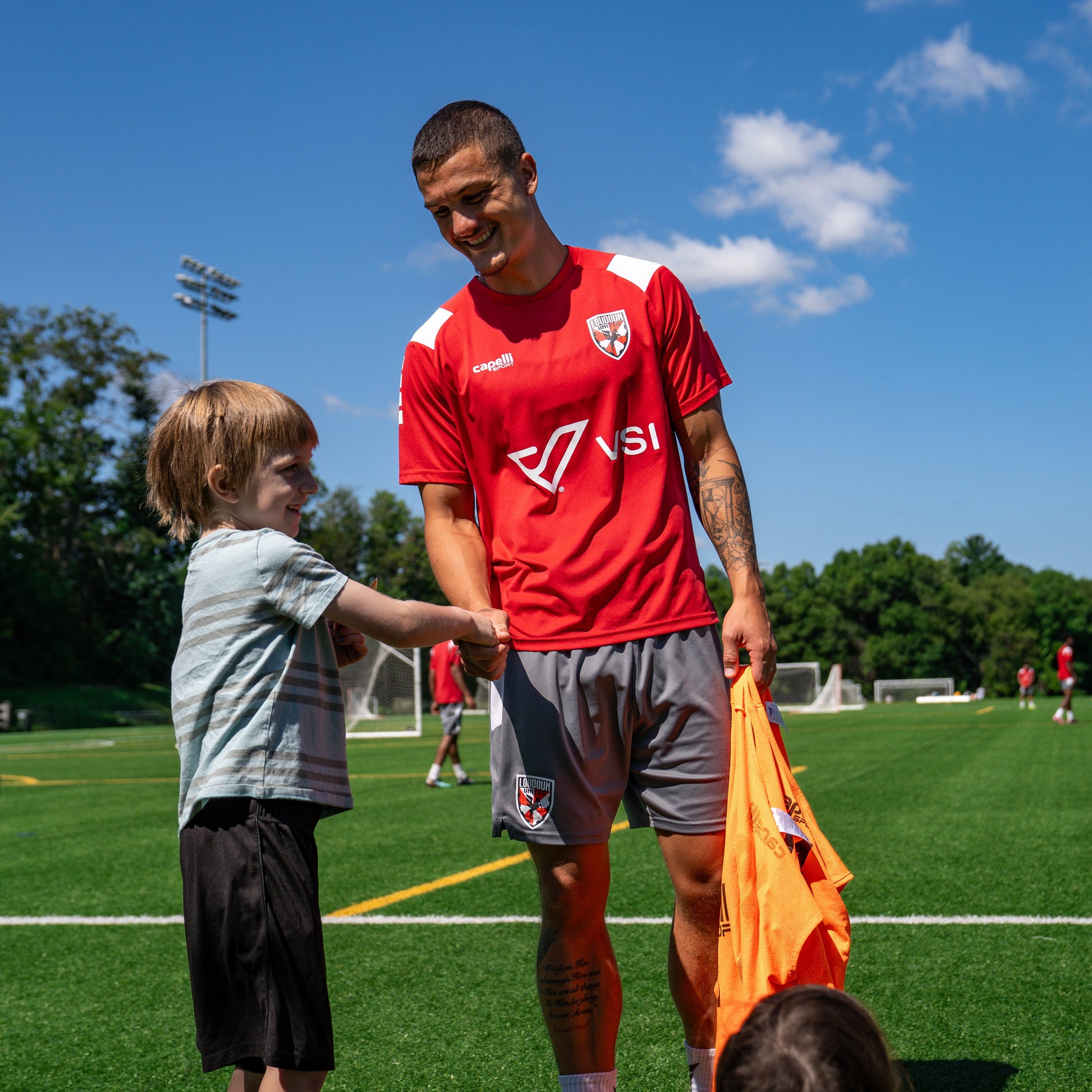 A soccer player in a Loudoun United FC red VSI training jersey shaking hands with a young child on a green soccer field .