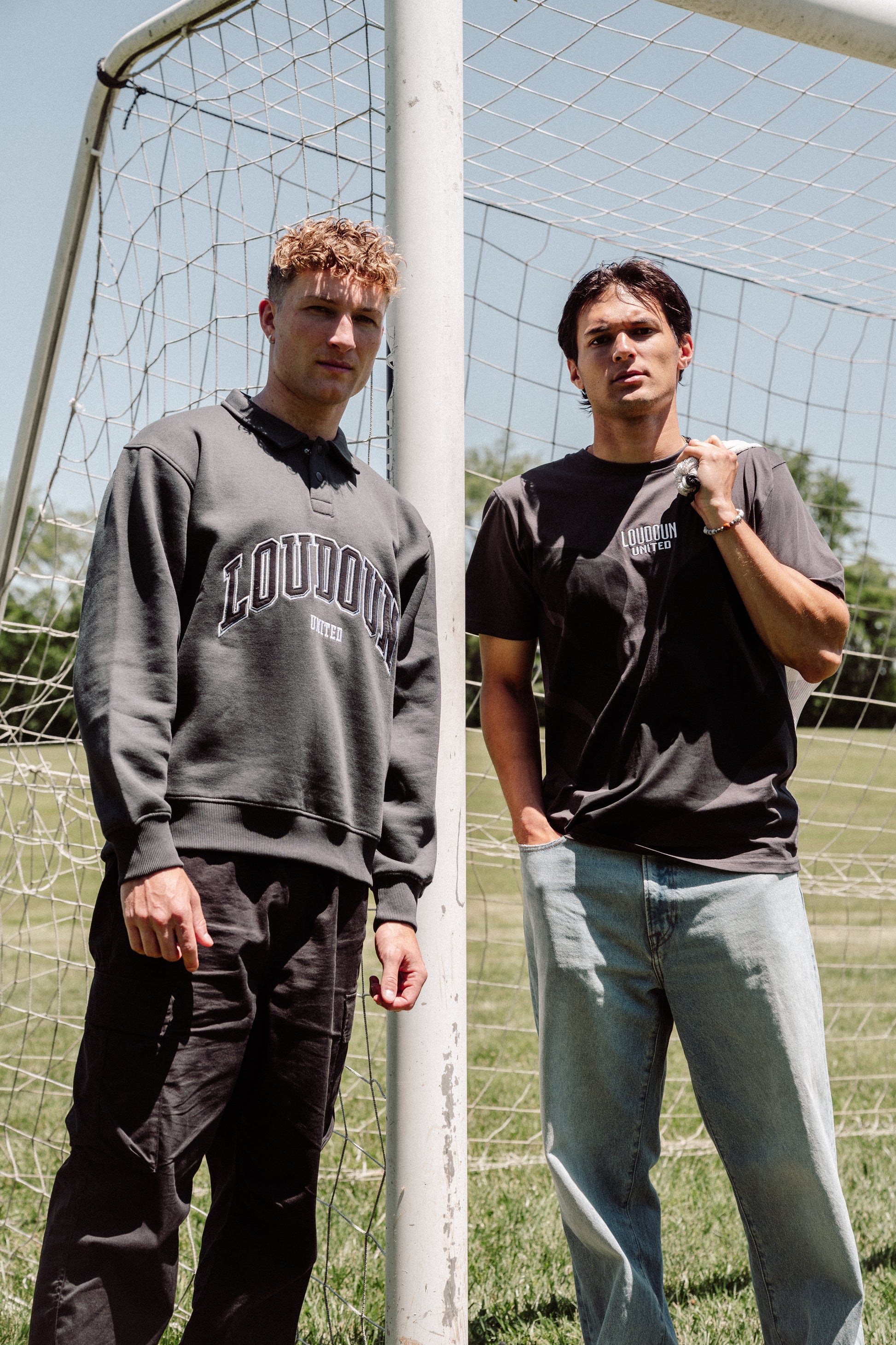 Two men standing in front of a soccer goal, one wearing a gray sweatshirt with 'LOUDON' and the other in a black t-shirt and jeans.