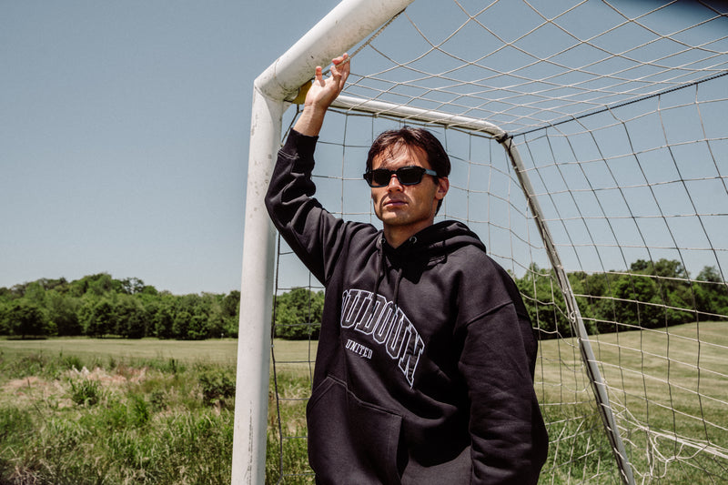Man wearing a black hoodie with text, standing next to a soccer goal on a field.
