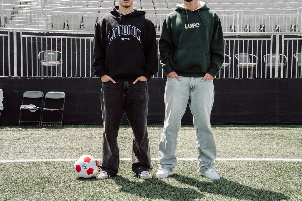 Two people standing on a soccer field with a soccer ball, wearing hoodies and jeans.