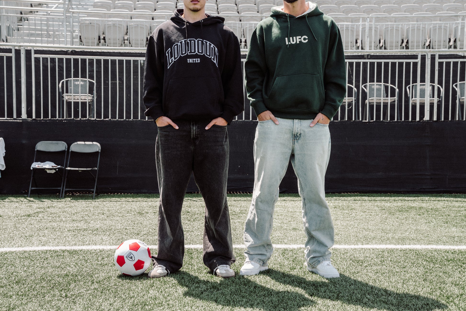 Two people standing on a soccer field with a soccer ball, wearing hoodies and jeans.